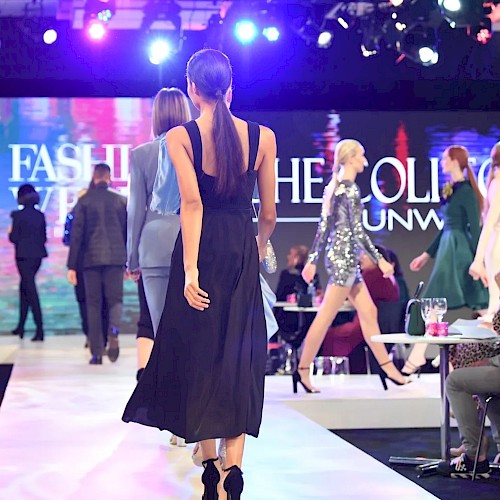 Models are walking down a runway during a fashion show with an audience seated on the sides. The background displays 