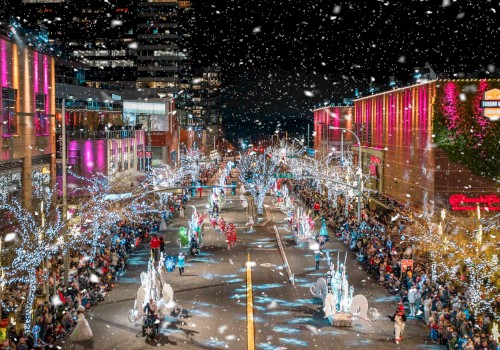 A festive street parade at night with colorful lights, decorated buildings, numerous participants, and spectators, in a snowy setting, is shown.