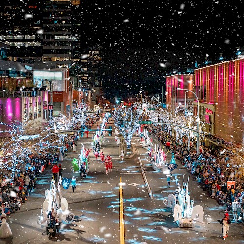 A festive street parade at night with colorful lights, decorated buildings, numerous participants, and spectators, in a snowy setting, is shown.