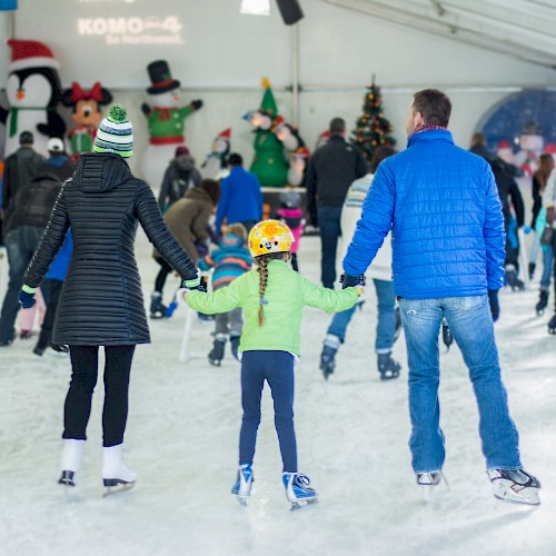 People are ice skating at an indoor rink, holding hands, while festive decorations adorn the background.