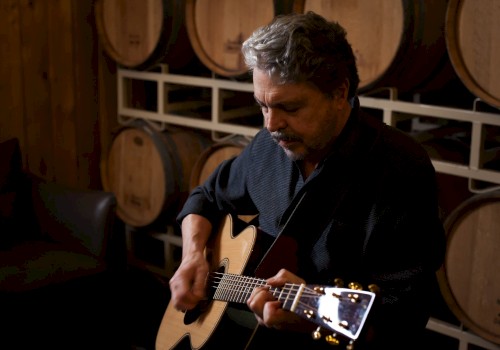 A man is playing an acoustic guitar in a room with wooden barrels stacked in the background, adding a rustic ambiance to the scene.