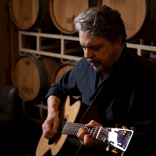 A man is playing an acoustic guitar in a room with wooden barrels stacked in the background, adding a rustic ambiance to the scene.