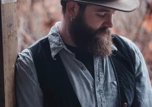 A bearded man in a hat and vest leans against a wooden post with a serene background of trees and nature.