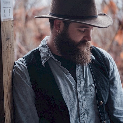 A bearded man in a hat and vest leans against a wooden post with a serene background of trees and nature.