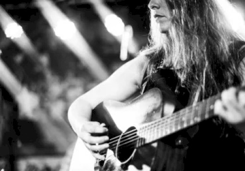 A black and white image shows a person playing an acoustic guitar on stage, illuminated by stage lights in the background.