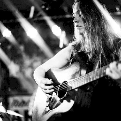 A black and white image shows a person playing an acoustic guitar on stage, illuminated by stage lights in the background.