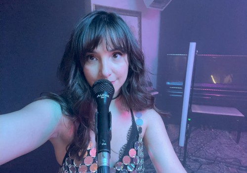 A woman with long hair and bangs is taking a selfie while singing into a microphone, standing in what appears to be a music or performance studio.