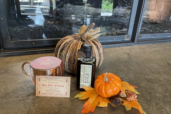 The image features a fall-themed arrangement with a pumpkin, leaves, a bottle, a tin, and a sign for wine tasting set in front of a fireplace.