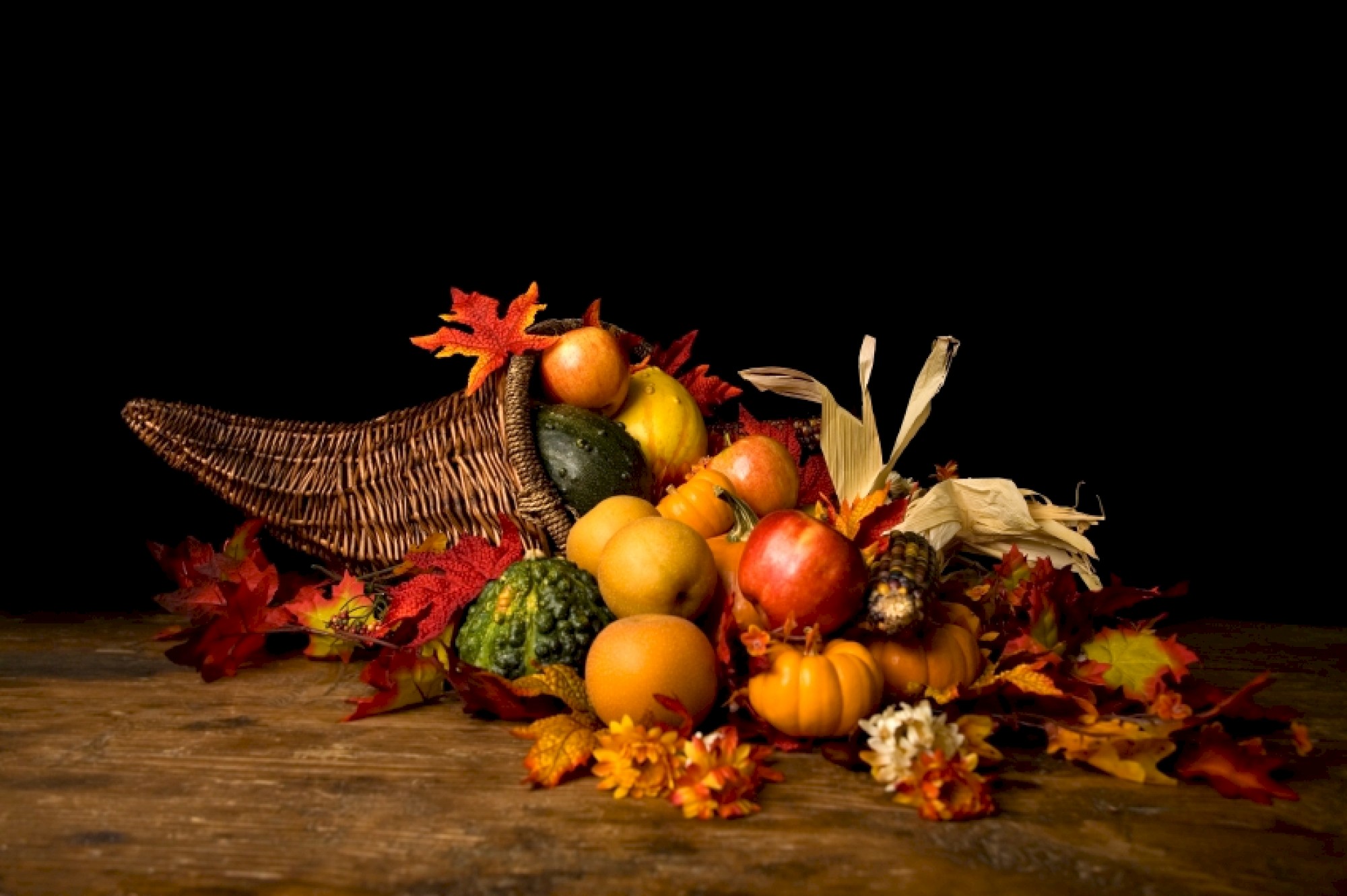 A cornucopia filled with various fruits and vegetables, surrounded by autumn leaves and flowers, sits on a wooden surface against a black background.