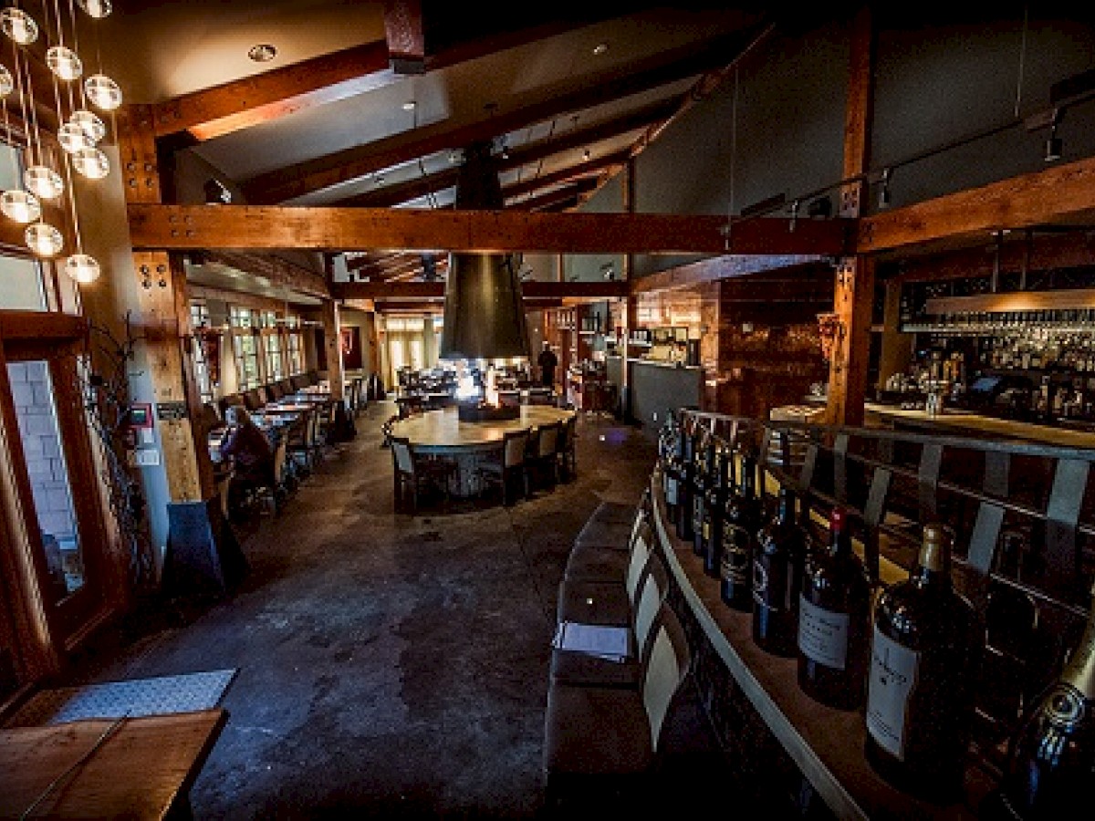 A rustic restaurant interior with a central circular seating area, wooden beams, a wine display along a counter, and ambient lighting from hanging bulbs.