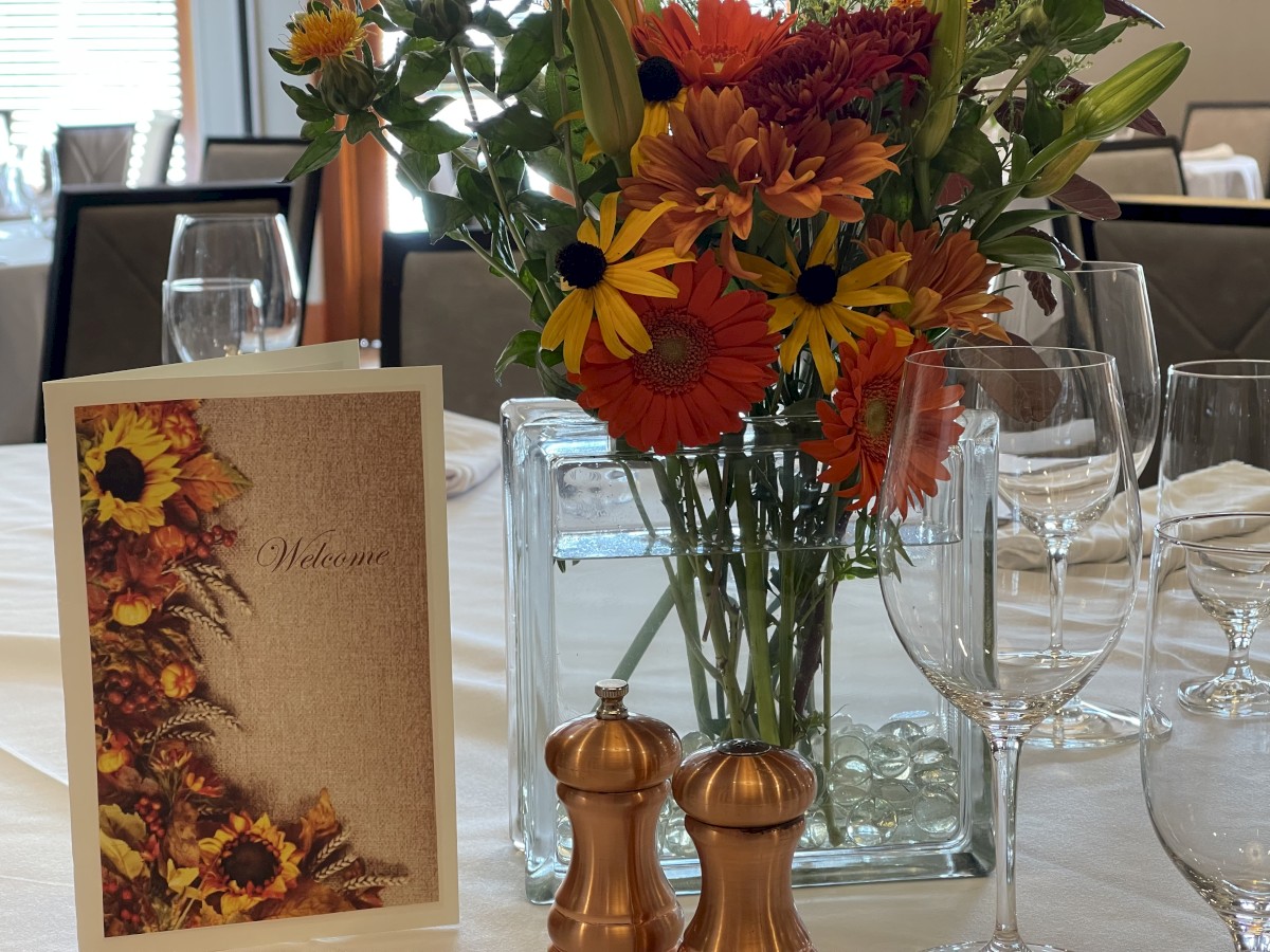 A table setting with a floral centerpiece, greeting card, wine glasses, and salt and pepper shakers on a white tablecloth.