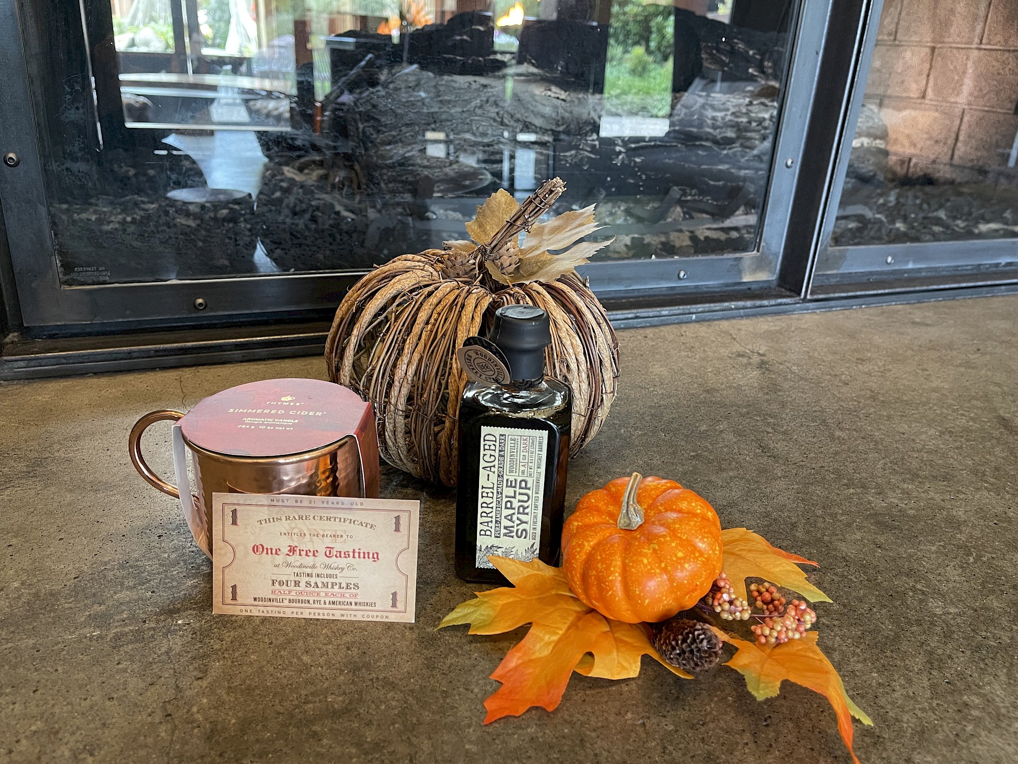 The image shows a fall-themed arrangement with a brown pumpkin, a bottle of olive oil, a mug, and a small orange pumpkin with leaves and pine cones.