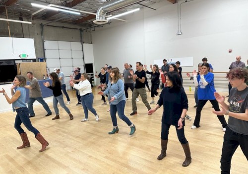 A group of people is participating in a dance class or practicing a routine in a spacious room with wooden floors and high ceilings.