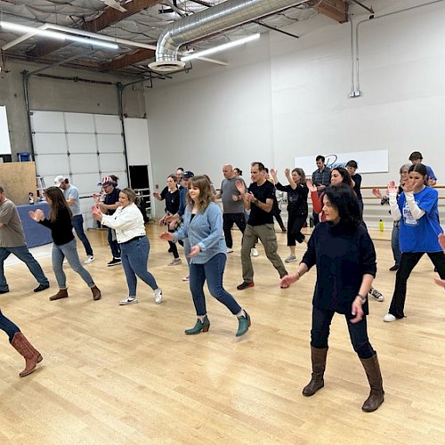 A group of people is participating in a dance class or practicing a routine in a spacious room with wooden floors and high ceilings.