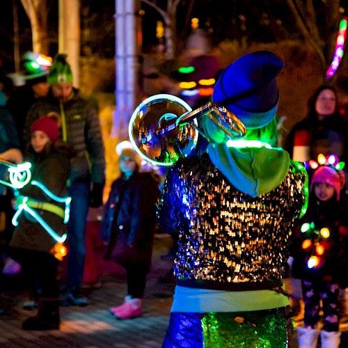 A person with glowing attire plays a trumpet outdoors, surrounded by a crowd in colorful, illuminated outfits.