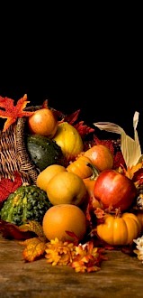 A cornucopia with assorted fruits, vegetables, and autumn leaves is displayed on a wooden surface against a black background.