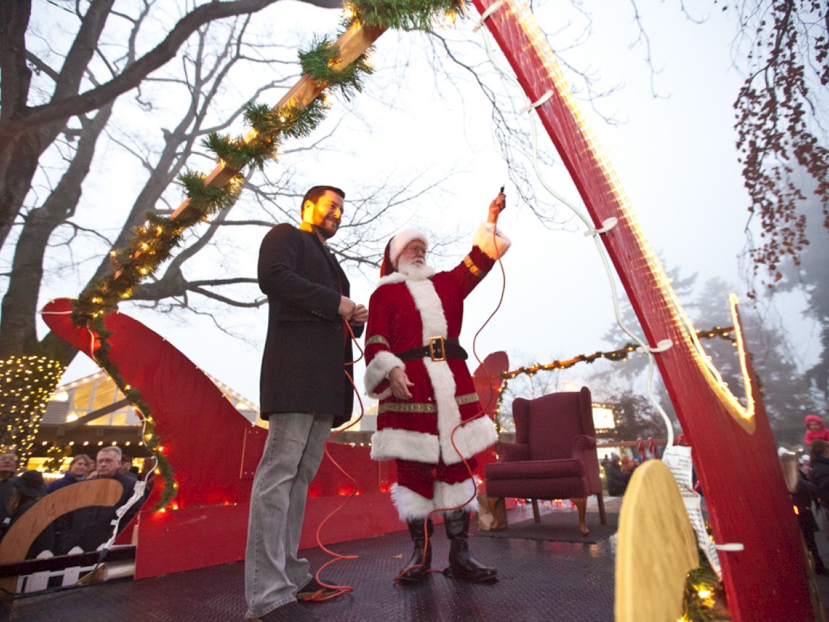 A person dressed as Santa Claus stands on a decorated float, holding a switch with another person, surrounded by festive lights and a crowd.