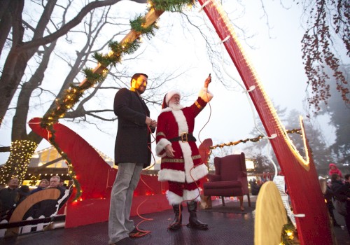 Santa in a red sleigh with a man, surrounded by Christmas lights and decorations, in front of a crowd.