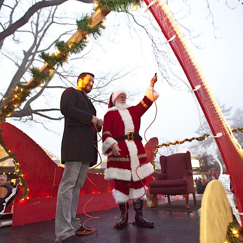 Santa in a red sleigh with a man, surrounded by Christmas lights and decorations, in front of a crowd.