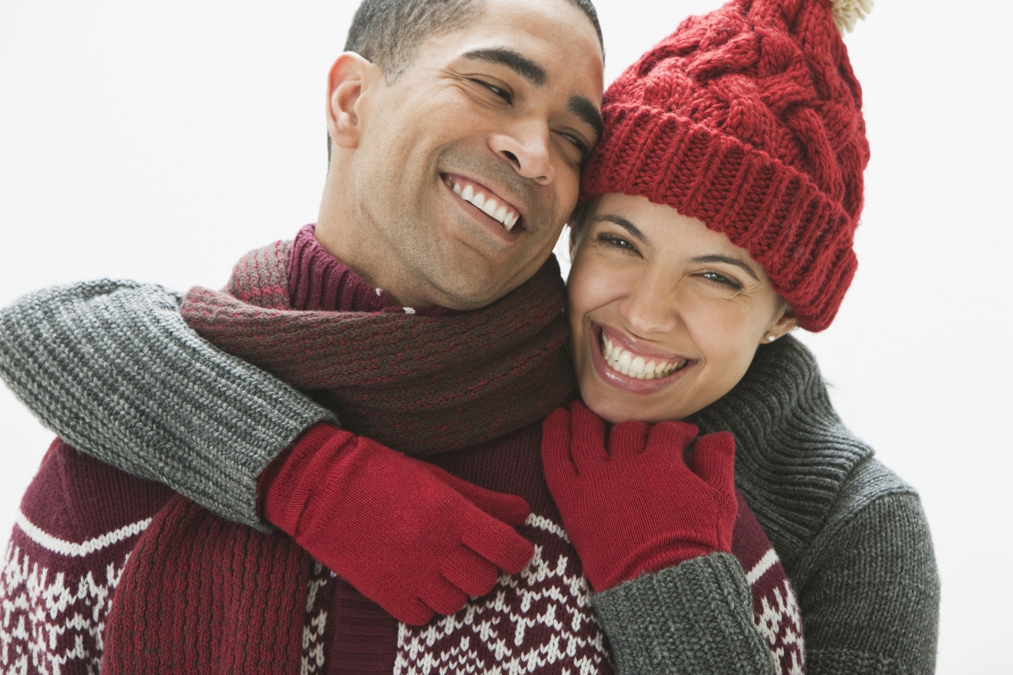 A couple is smiling and embracing each other, dressed warmly in winter clothes including hats, scarves, and gloves.