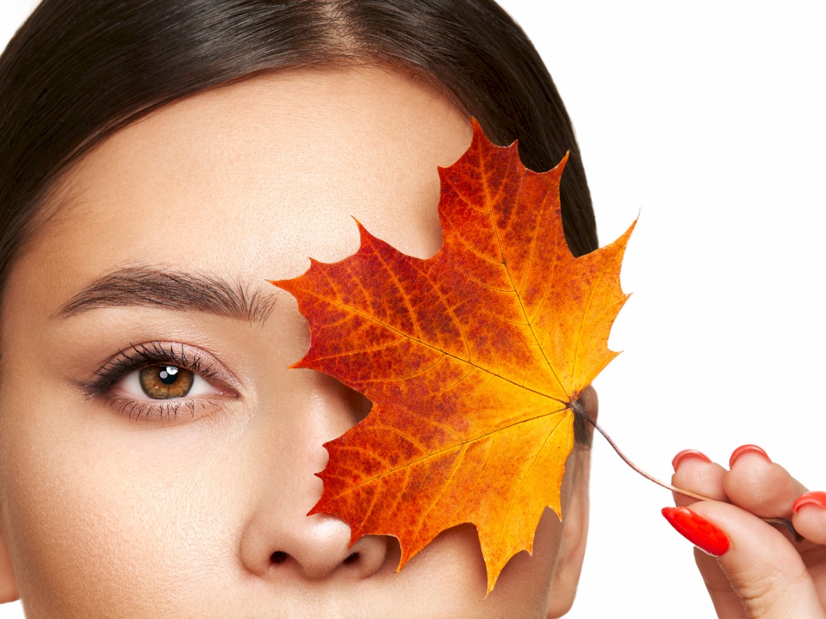 A person with dark hair holds an orange maple leaf over one eye, with a neutral expression against a white background.