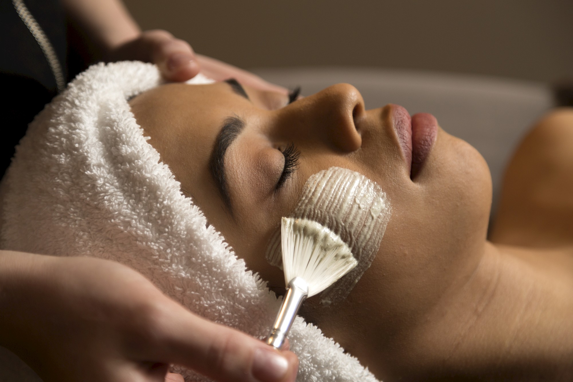 A person receiving a facial treatment with a towel around their head; a brush is applying a facial mask.