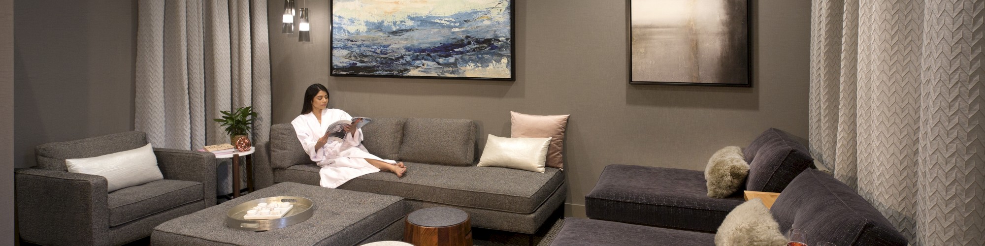 A modern living room with gray sofas, two paintings on the wall, and a person reading. There's a cozy vibe with warm lighting.