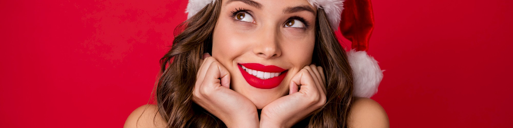 A person wearing a Santa hat is smiling and looking upward against a red background.