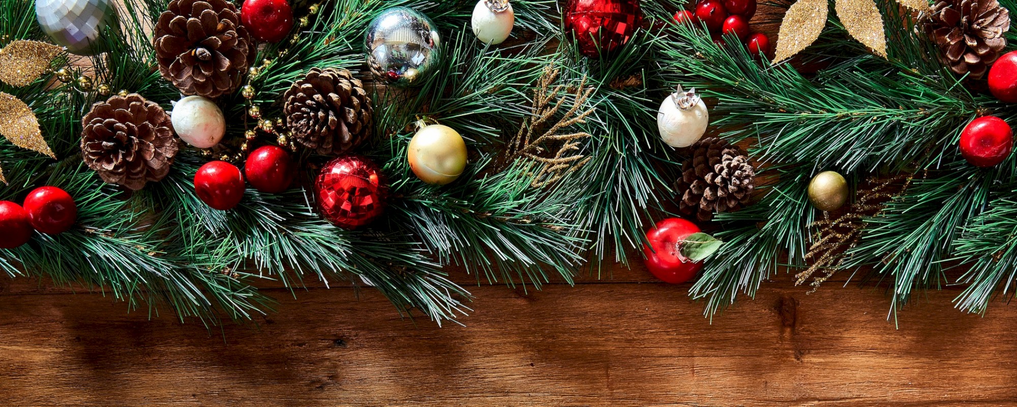 The image shows a festive Christmas garland with pine cones, red and gold ornaments, and green foliage on a wooden surface.