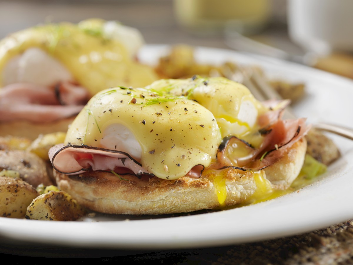 The image shows eggs Benedict with ham on toasted English muffins, topped with hollandaise sauce, served alongside breakfast potatoes.