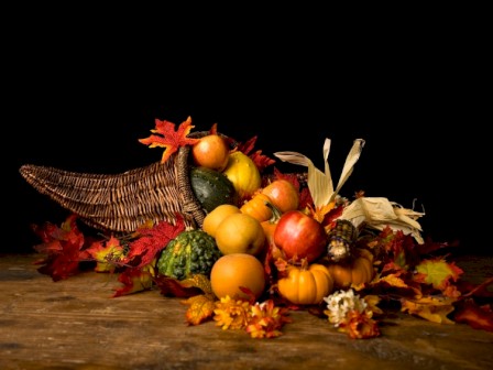 A cornucopia overflowing with autumn harvest fruits and vegetables, surrounded by colorful fall leaves, on a rustic wooden surface.