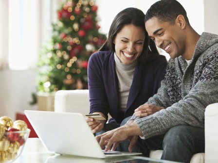 A couple is smiling while using a laptop together in a festive living room with a Christmas tree in the background.