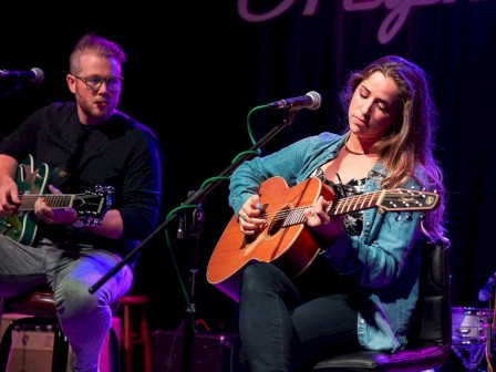 Two musicians are playing guitars on stage, each in front of a microphone, with a dimly lit backdrop visible behind them.