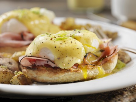 Eggs Benedict with ham on an English muffin, topped with hollandaise sauce, garnished with herbs, served with potatoes.