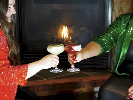 Two people in festive attire are toasting with drinks in front of a fireplace.