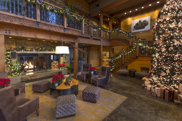 A cozy room with Christmas decor, a large tree, presents, and a fireplace. Comfortable seating and a staircase add a warm, inviting touch.