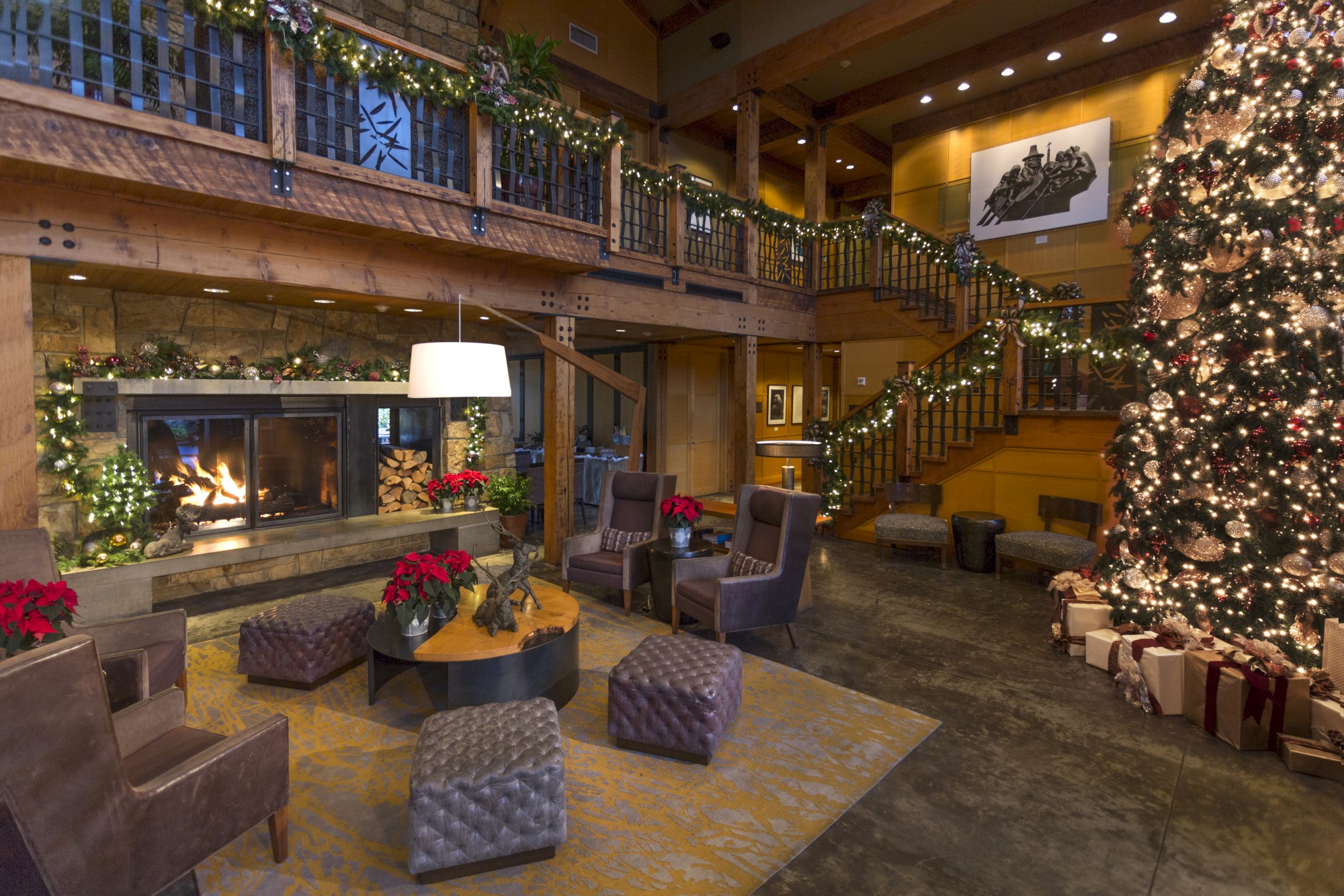 A cozy, decorated room with a large Christmas tree, fireplace, chairs, and festive garlands on a staircase.