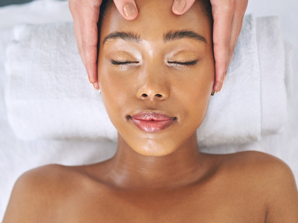 A person is lying on a massage table with eyes closed, covered with a white towel, receiving a head massage.