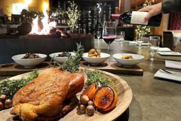 A roast bird with herbs and citrus on a platter, surrounded by side dishes, as wine is poured into a glass in a cozy setting.