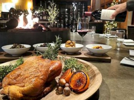 A roasted chicken with herbs and oranges is served on a wooden platter. Wine is being poured, and a cozy restaurant setting surrounds it.