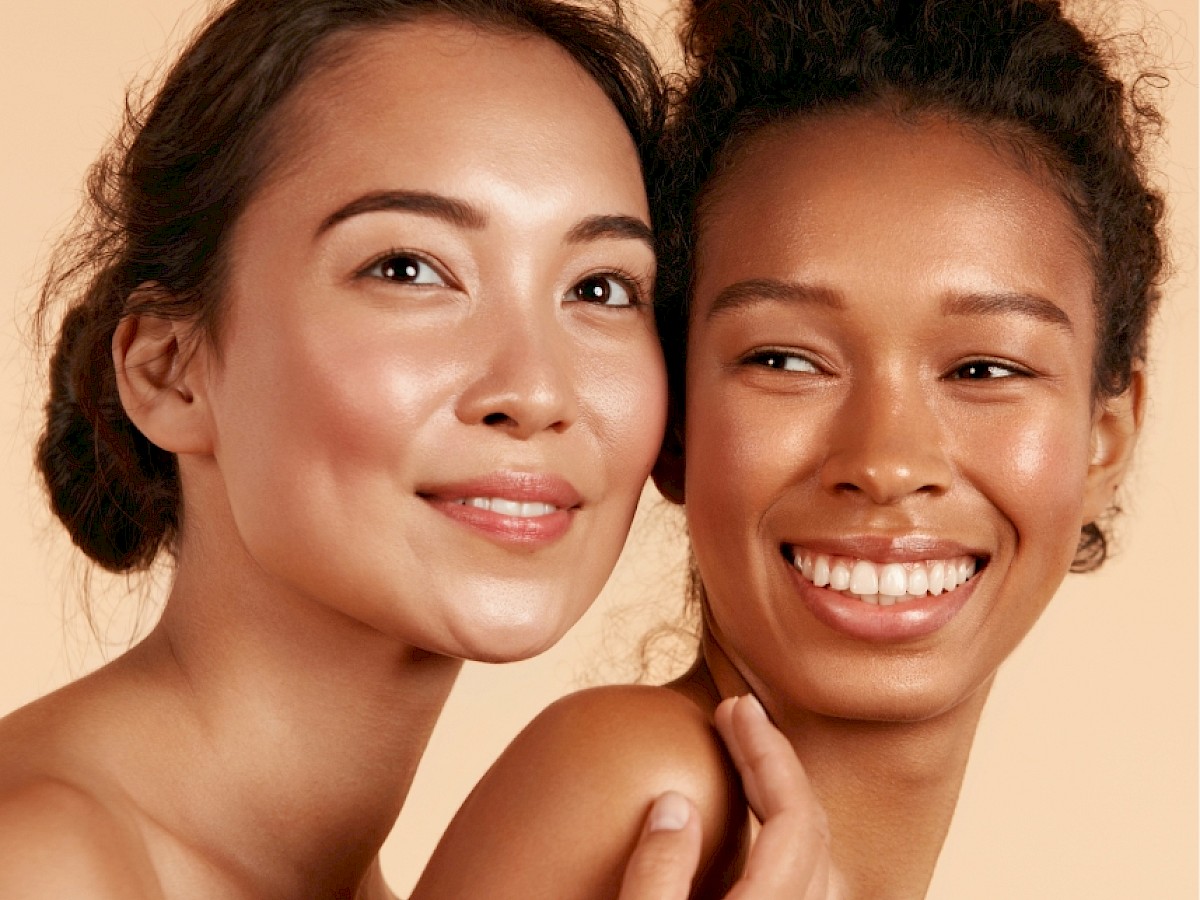 Two women smiling, close together against a light background, showcasing natural beauty with radiant skin and effortless hairstyles.