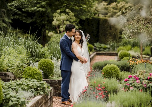 A couple in wedding attire embraces in a lush garden surrounded by greenery and flowers.