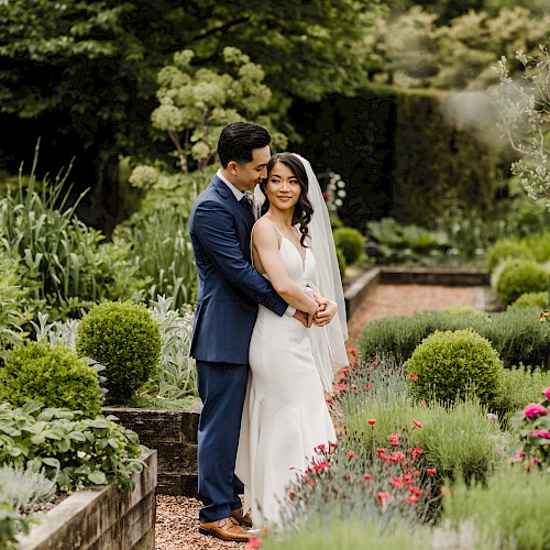 A couple in wedding attire embraces in a lush garden surrounded by greenery and flowers.