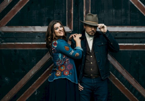 A woman and man stand in front of a wooden door; she smiles, and he adjusts his hat, both in stylish, layered outfits.