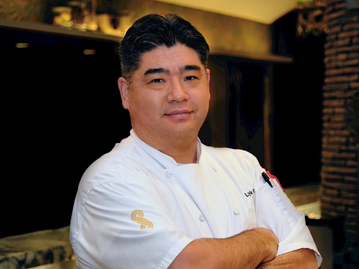 A person in a white chef's coat stands with arms crossed in a kitchen or restaurant setting.