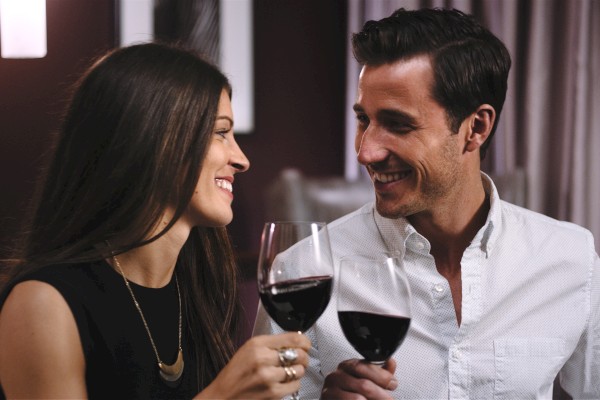 A couple is smiling and toasting with glasses of red wine, seated in a cozy indoor setting.