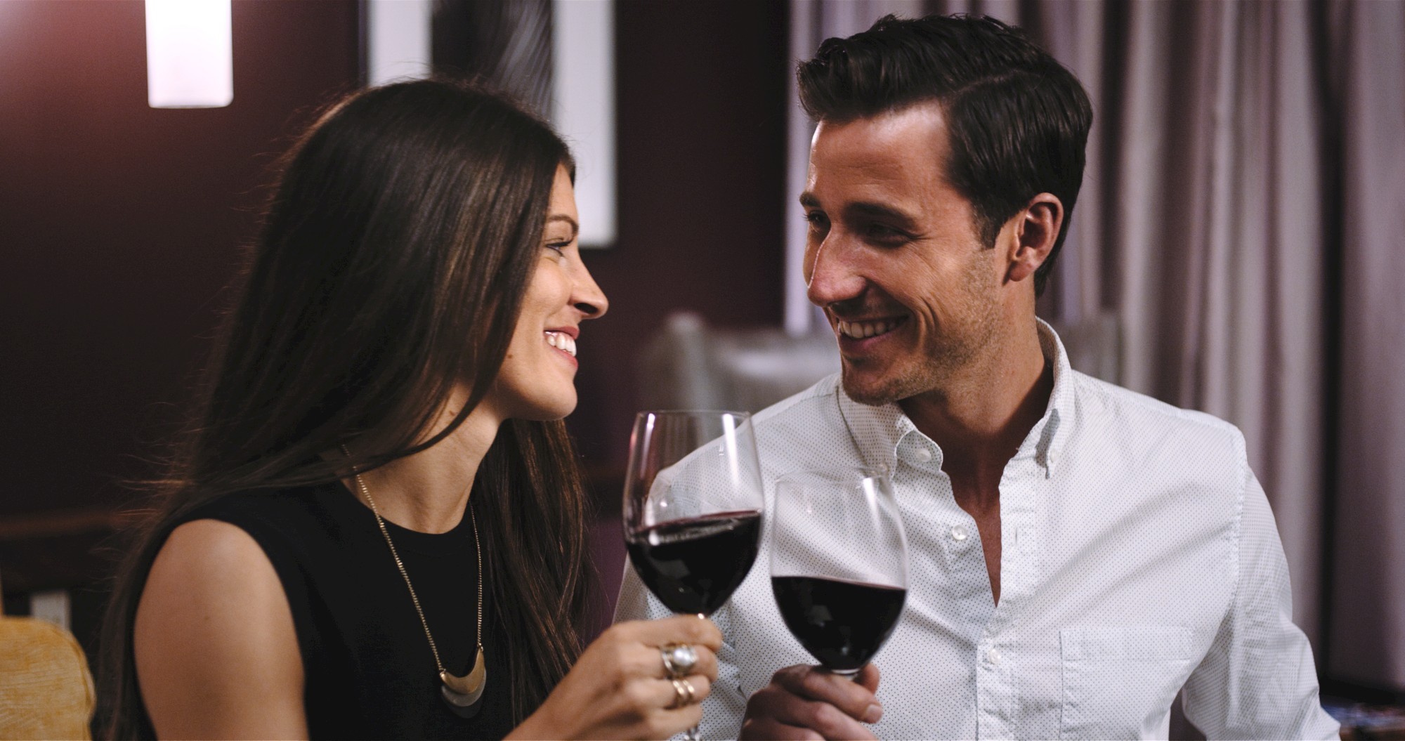 A man and woman are smiling and clinking glasses of red wine in a cozy indoor setting.