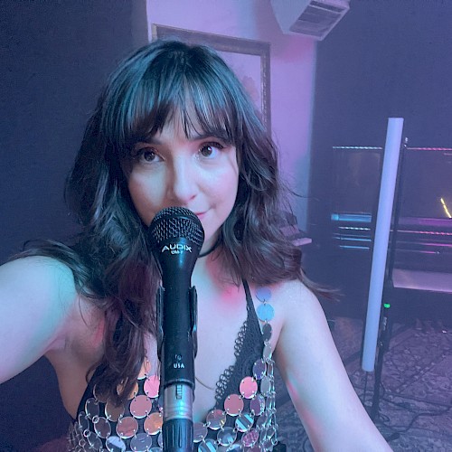 A person taking a selfie with a microphone, wearing a shiny top, possibly in a music or performance setting with dim lighting.