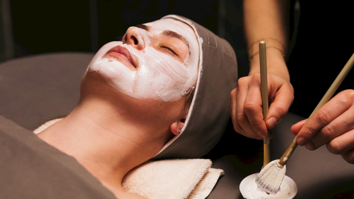 A person is lying down with a facial mask applied. Another individual is using a brush to apply or adjust the mask.