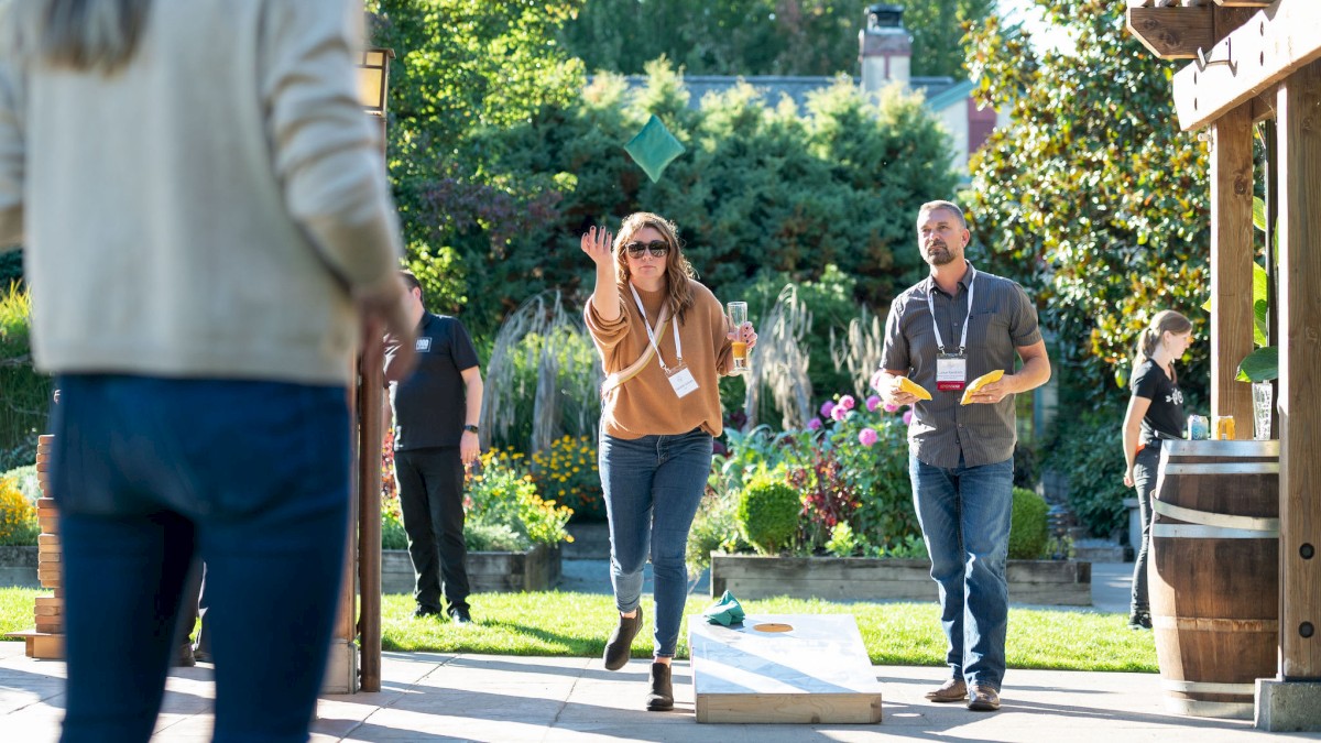 People are playing cornhole outdoors, with one person tossing a bean bag. Others watch and hold drinks, enjoying the sunny setting.